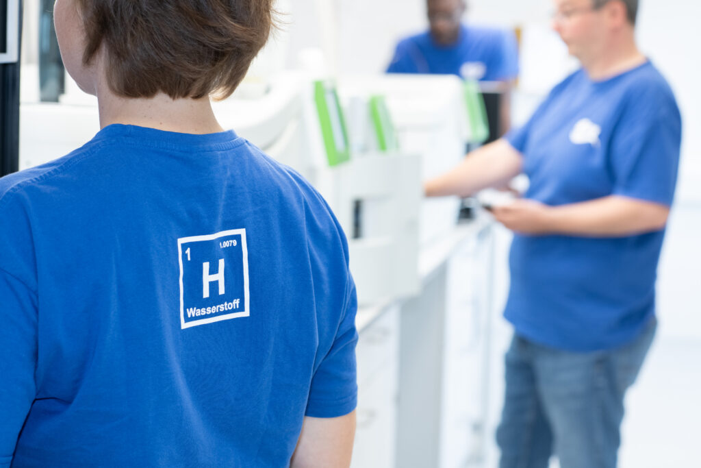 Drei Mitarbeiter in blauen T-Shirts stehen an H2-Analytik-Anlagen